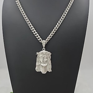 Puede incluir: Collar plateado con un colgante de Jesús con una corona de espinas. El colgante cuelga de una gruesa cadena de eslabones. El collar se exhibe en un soporte negro, mostrando la joyería religiosa.
