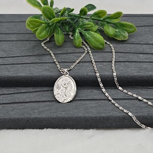 Puede incluir: Un collar con colgante ovalado plateado con una imagen grabada de un santo. El colgante cuelga de una delicada cadena. El collar se exhibe sobre una superficie gris oscuro, con follaje verde en el fondo.