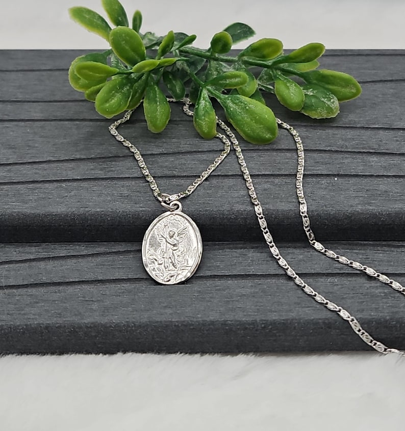Puede incluir: Un collar con colgante ovalado plateado con una imagen grabada de un santo. El colgante cuelga de una delicada cadena. El collar se exhibe sobre una superficie gris oscuro, con follaje verde en el fondo.