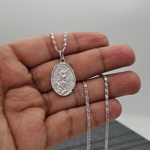 Puede incluir: Un collar con colgante ovalado plateado con un relieve detallado de una figura alada, posiblemente un &aacute;ngel, sobre una cadena. El colgante cuelga de una fina cadena, el dise&ntilde;o es visible sobre un fondo neutro.