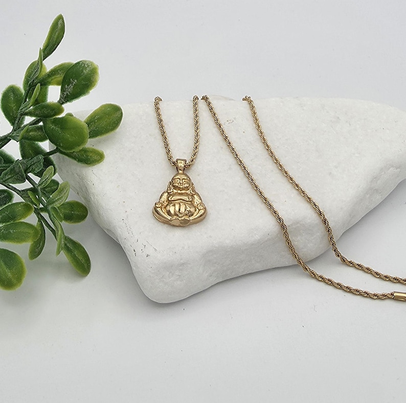 May include: A gold-toned necklace featuring a pendant shaped like a seated, smiling Buddha figure. The pendant is attached to a thin, twisted rope chain. The metal has a polished finish, reflecting light across the surface of the figure.