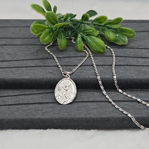 Puede incluir: Un collar plateado con un colgante ovalado que representa un &aacute;ngel. El colgante cuelga de una fina cadena. Hay follaje verde en el fondo, sobre una superficie gris oscuro.