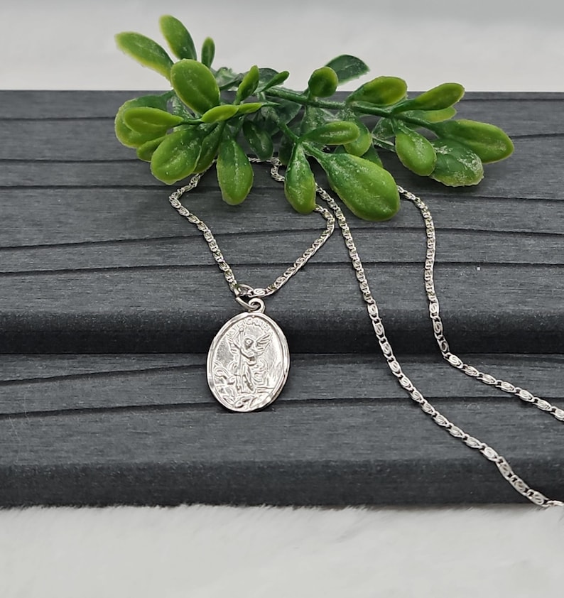 Puede incluir: Un collar plateado con un colgante ovalado que representa un &aacute;ngel. El colgante cuelga de una fina cadena. Hay follaje verde en el fondo, sobre una superficie gris oscuro.