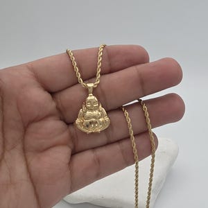 May include: A gold-toned pendant shaped like a seated figure with a smooth, rounded head and body, suspended from a matching gold-toned rope chain. The pendant features a polished metallic finish that reflects light across its curved surface.