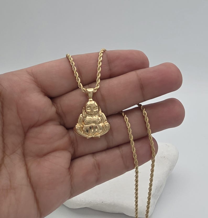 May include: A gold-toned pendant shaped like a seated figure with a smooth, rounded head and body, suspended from a matching gold-toned rope chain. The pendant features a polished metallic finish that reflects light across its curved surface.