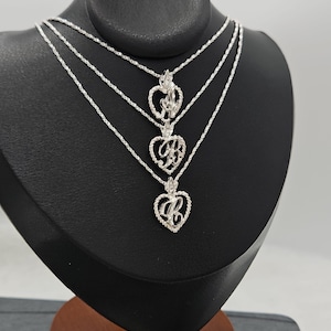 May include: Three silver-toned necklaces featuring heart-shaped pendants with an intricate, textured border. Each pendant contains a stylised, cursive letter in the centre. The necklaces are layered, with each chain having a delicate, twisted link design.