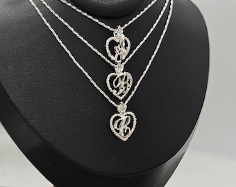 Puede incluir: Tres collares de tono plateado con colgantes en forma de corazón que presentan un borde texturizado intrincado. Cada colgante contiene una letra estilizada en cursiva en el centro. Los collares están superpuestos con cadenas de eslabones trenzados.