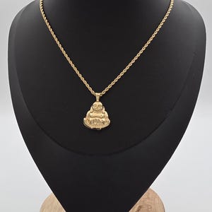 May include: A gold-toned rope chain necklace featuring a pendant shaped like a seated, smiling Buddha figure. The pendant is polished and reflective, suspended from a twisted metal chain that rests against a black display form.