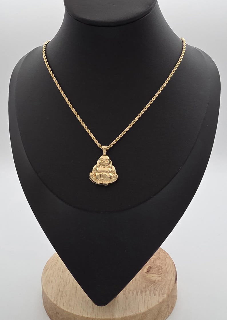 May include: A gold-toned rope chain necklace featuring a pendant shaped like a seated, smiling Buddha figure. The pendant is polished and reflective, suspended from a twisted metal chain that rests against a black display form.