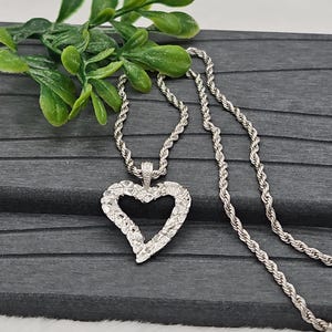 Puede incluir: Un collar plateado con un colgante en forma de corazón. El colgante tiene una superficie texturizada y una pequeña piedra transparente en la parte superior. La cadena es de diseño trenzado. El collar se muestra sobre una superficie gris oscuro con follaje verde.