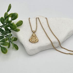 May include: A gold-toned necklace featuring a pendant shaped like a seated Buddha figure. The pendant is attached to a thin, twisted rope chain. The metal has a polished finish, reflecting light across the surface of the figure and the chain links.
