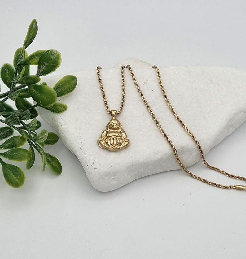 May include: A gold-toned necklace featuring a pendant shaped like a seated Buddha figure. The pendant is attached to a thin, twisted rope chain. The metal has a polished finish, reflecting light across the surface of the figure and the chain links.