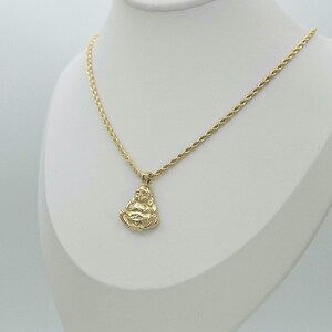 May include: A gold-colored necklace featuring a rope chain and a pendant shaped like a seated figure in a meditative pose. The pendant has a polished finish and is suspended from the center of the chain, which rests on a white display form.