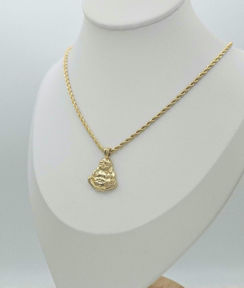 May include: A gold-colored necklace featuring a rope chain and a pendant shaped like a seated figure in a meditative pose. The pendant has a polished finish and is suspended from the center of the chain, which rests on a white display form.