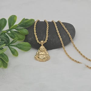 May include: A gold-toned metal pendant shaped like a seated Buddha figure, suspended from a matching gold-toned rope chain. The pendant features a polished finish with detailed facial features and a rounded belly, reflecting light across its surface.