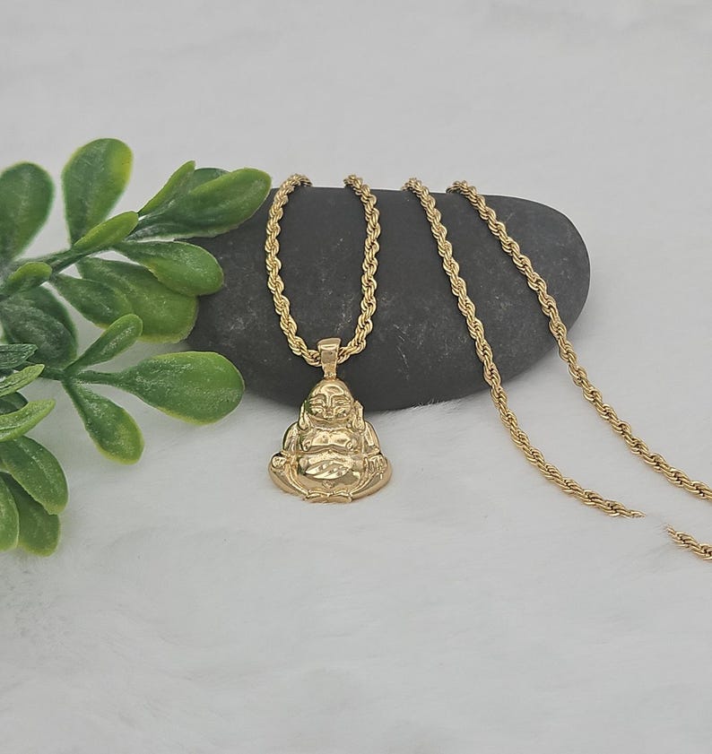 May include: A gold-toned metal pendant shaped like a seated Buddha figure, suspended from a matching gold-toned rope chain. The pendant features a polished finish with detailed facial features and a rounded belly, reflecting light across its surface.