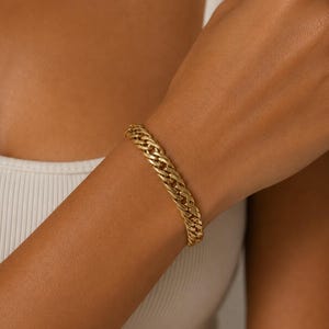 May include: A gold-toned chain link bracelet featuring a series of interlocking, polished metal loops. The bracelet has a substantial, textured appearance with a repeating pattern of connected links that wrap around the wrist in a continuous design.
