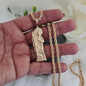 May include: Gold-coloured pendant necklace featuring a detailed figure with a halo, holding a staff. The pendant hangs from a rope chain. The necklace is displayed on a white surface.