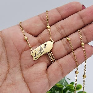 May include: Gold necklace with a pendant shaped like the island of Puerto Rico. The pendant has the name "Lydia" and the year "1939" engraved on it. The necklace has small gold beads along the chain.