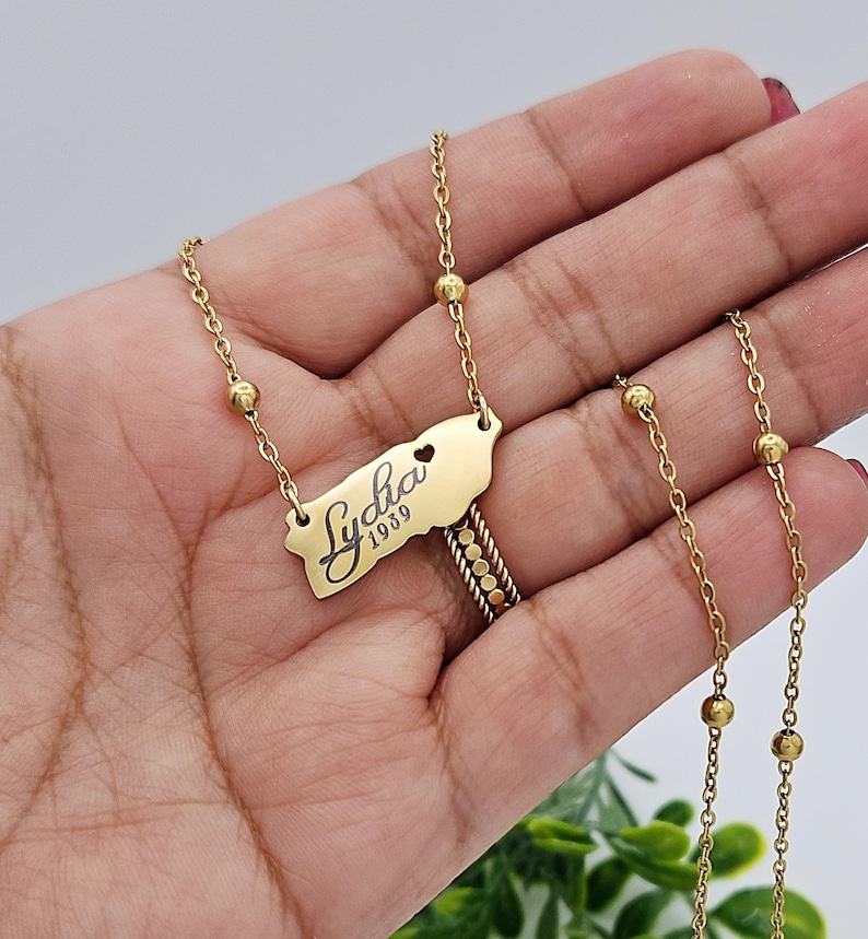 May include: Gold necklace with a pendant shaped like the island of Puerto Rico. The pendant has the name "Lydia" and the year "1939" engraved on it. The necklace has small gold beads along the chain.