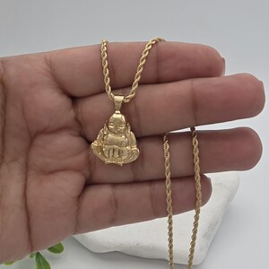 May include: A gold-toned rope chain necklace featuring a pendant shaped like a seated figure with hands resting on the lap. The pendant has a smooth, polished finish and is suspended from a twisted metal chain that drapes down the length of the piece.