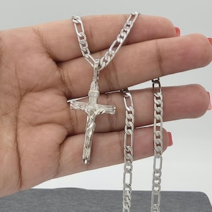 May include: A silver-colored crucifix pendant necklace. The pendant features a detailed depiction of Jesus on the cross. The chain is a curb link style. The necklace is displayed against a neutral background.