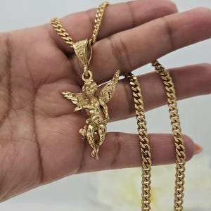 K&ouml;nnte beinhalten: Goldfarbene Halskette mit detailliertem Cherub-Anh&auml;nger und strukturierter Kette. Der Anh&auml;nger zeigt einen Cherub mit ausgebreiteten Fl&uuml;geln. Kette und Anh&auml;nger haben eine polierte Oberfl&auml;che, die das Licht reflektiert. Ein klassisches Schmuckst&uuml;ck.