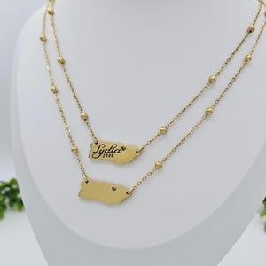 May include: Gold layered necklace with two pendants. The top pendant is a gold outline of Puerto Rico with the name "Lydia" and the year "1989" inscribed. The bottom pendant is a gold outline of Puerto Rico with a small heart in the center.
