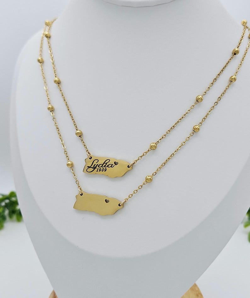 May include: Gold layered necklace with two pendants. The top pendant is a gold outline of Puerto Rico with the name "Lydia" and the year "1989" inscribed. The bottom pendant is a gold outline of Puerto Rico with a small heart in the center.