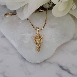 Puede incluir: Un collar de cadena de oro con un peque&ntilde;o colgante de &aacute;ngel. El &aacute;ngel est&aacute; representado con alas y un halo.