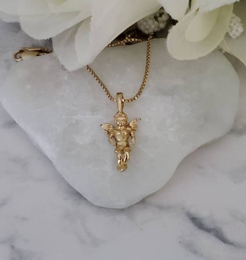 Puede incluir: Un collar de cadena de oro con un peque&ntilde;o colgante de &aacute;ngel. El &aacute;ngel est&aacute; representado con alas y un halo.