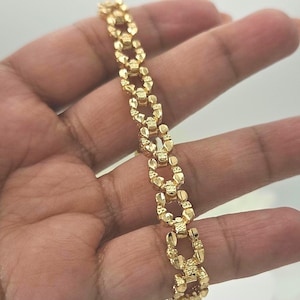 Könnte beinhalten: Ein goldfarbenes Armband mit einem sich wiederholenden Muster aus verbundenen, strukturierten Formen. Das Armband wird in einer Hand gehalten, um sein Design und seine Farbe zu zeigen. Das Schmuckstück scheint aus Metall zu sein.