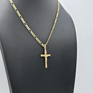 Puede incluir: Un collar de cadena de oro con un colgante de cruz de oro. La cadena es una cadena de estilo figaro.
