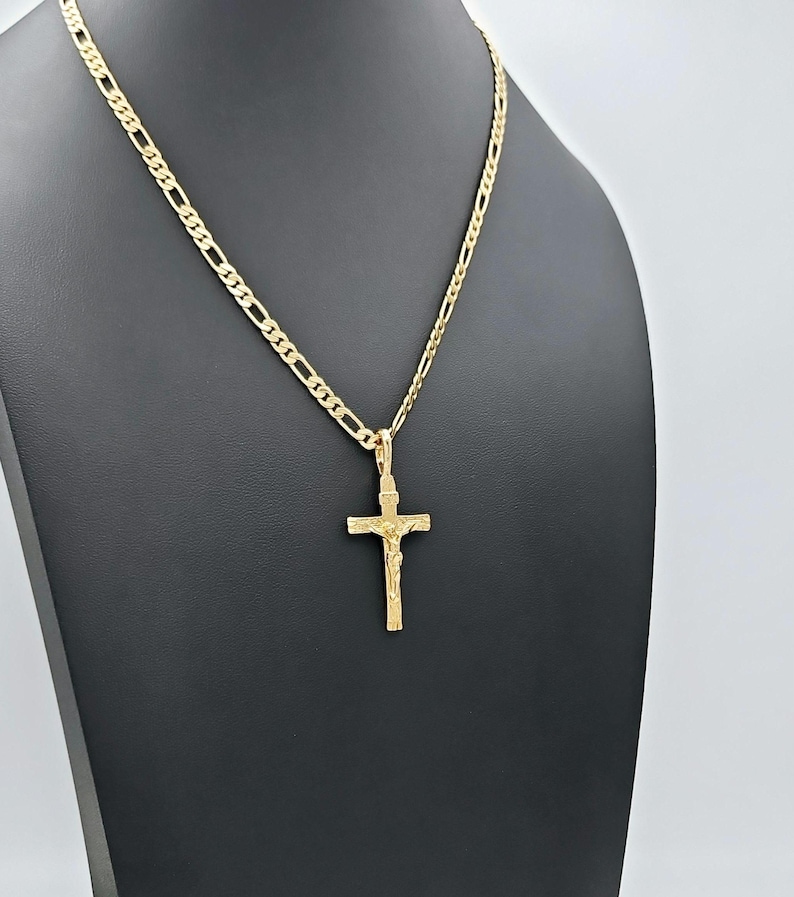 Puede incluir: Un collar de cadena de oro con un colgante de cruz de oro. La cadena es una cadena de estilo figaro.