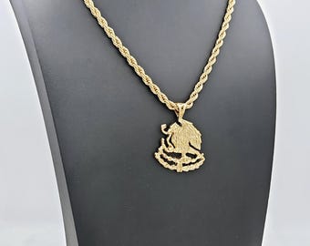May include: A gold rope chain necklace with a gold pendant featuring a detailed eagle design. The pendant is a stylized representation of the Mexican coat of arms.