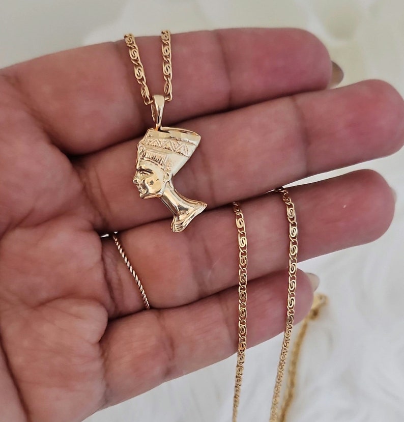 May include: Gold chain necklace with a pendant featuring a profile of an Egyptian queen wearing a headdress.