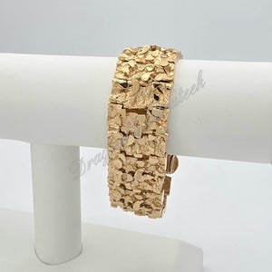May include: A gold bracelet with a textured, nugget-like design. The bracelet is displayed on a white stand, highlighting its intricate details and the warm tones of the metal. The clasp is visible, indicating its functionality.