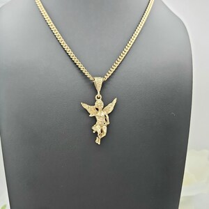 K&ouml;nnte beinhalten: Eine goldfarbene Halskette mit einem detaillierten Engel-Anh&auml;nger. Der Engel hat ausgebreitete Fl&uuml;gel und h&auml;ngt an einer Panzerkette. Die Halskette wird auf einer schwarzen Schaufensterpuppe pr&auml;sentiert, die das Design und die Farbe des Schmucks hervorhebt.