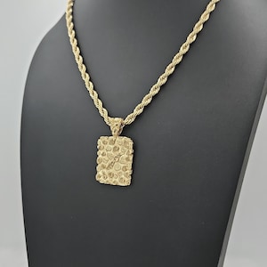 May include: A gold-colored necklace with a textured rectangular pendant. The chain is a twisted rope design. The pendant has a rough, uneven surface. The necklace is displayed on a black mannequin.
