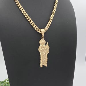 May include: Gold-coloured necklace with a pendant depicting a religious figure. The pendant hangs from a thick, textured chain. The figure has a halo and staff. The necklace is shown against a black backdrop.