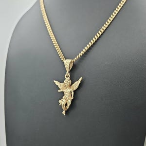 K&ouml;nnte beinhalten: Eine goldfarbene Halskette mit Anh&auml;nger, die einen detaillierten Engel-Anh&auml;nger zeigt. Der Engel hat ausgebreitete Fl&uuml;gel und h&auml;ngt an einer Panzerkette. Die Halskette wird vor einem dunklen Hintergrund pr&auml;sentiert.