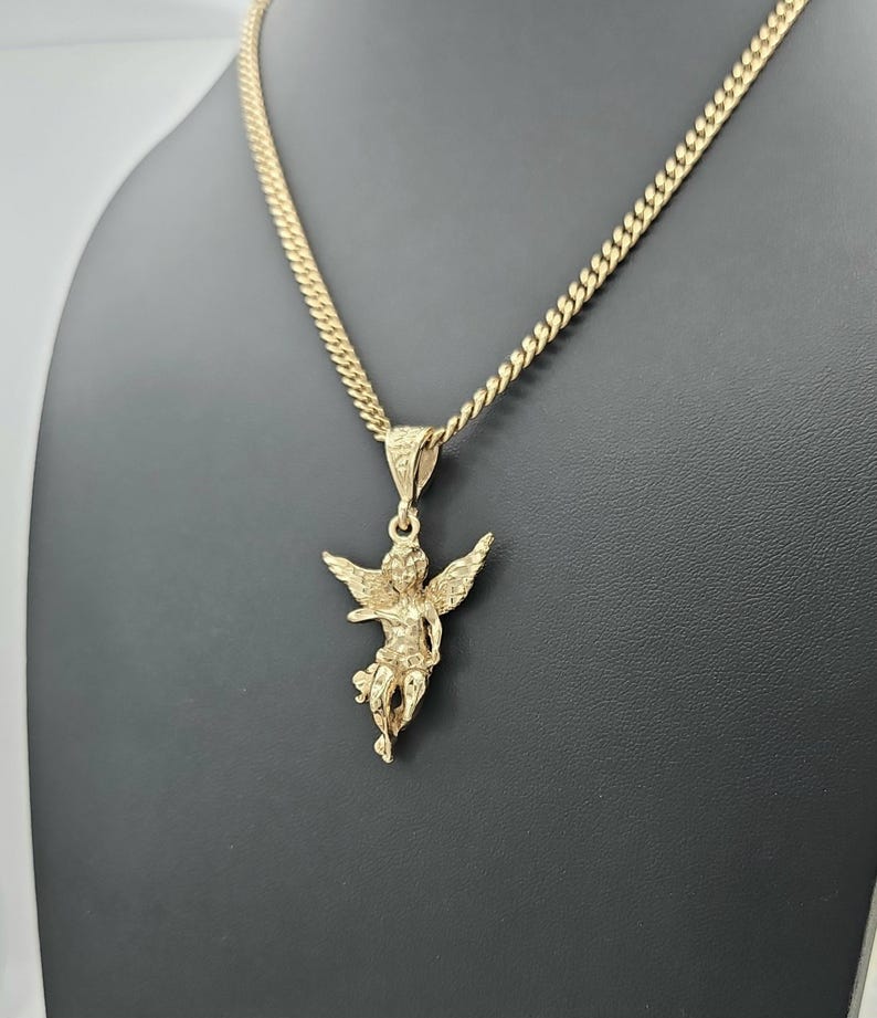 K&ouml;nnte beinhalten: Eine goldfarbene Halskette mit Anh&auml;nger, die einen detaillierten Engel-Anh&auml;nger zeigt. Der Engel hat ausgebreitete Fl&uuml;gel und h&auml;ngt an einer Panzerkette. Die Halskette wird vor einem dunklen Hintergrund pr&auml;sentiert.