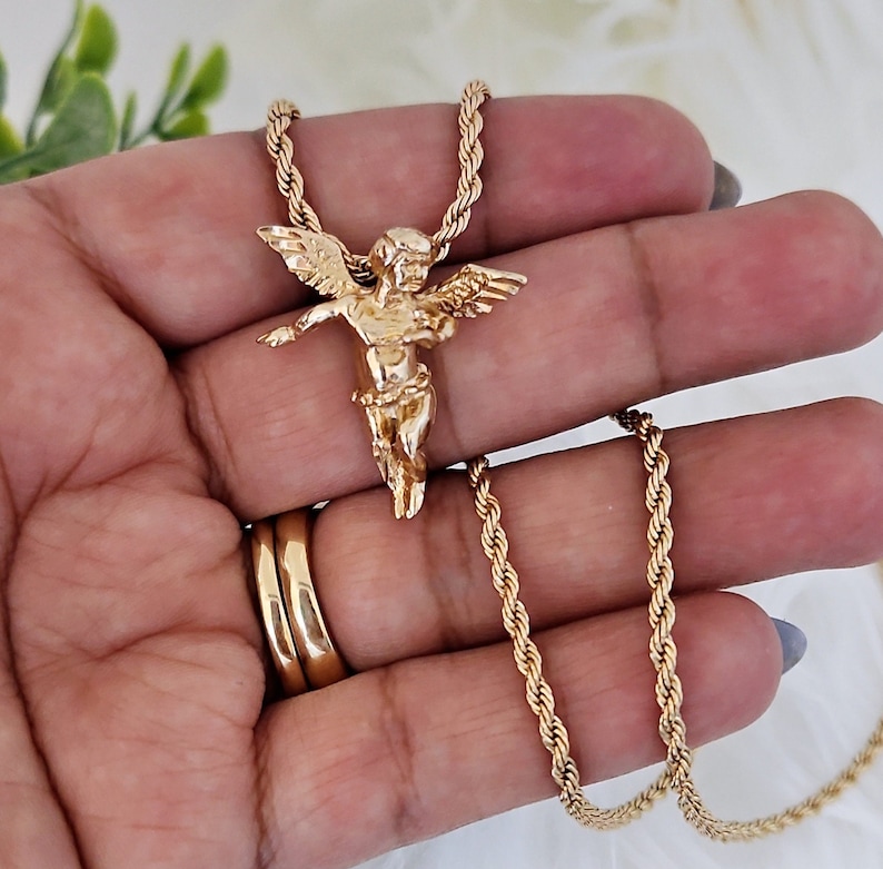 May include: Gold chain necklace with a gold cherub pendant. The chain is a rope style and the pendant is detailed.