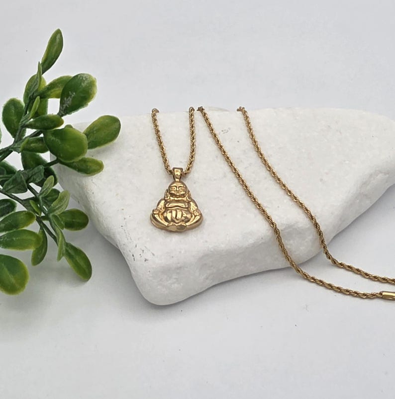 May include: A gold-toned pendant necklace featuring a seated Buddha figure with a smiling expression. The pendant is attached to a thin, twisted rope chain. The metal has a polished finish, reflecting light across the surface of the figure.