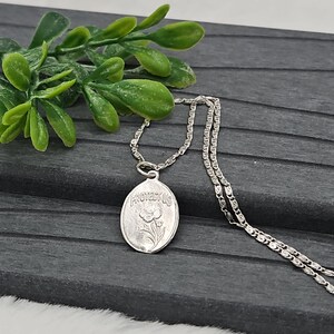Puede incluir: Collar plateado con un colgante ovalado con un dise&ntilde;o floral y la palabra "PROTECTUS". El colgante cuelga de una delicada cadena. Hojas verdes en el fondo.