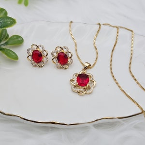 May include: A gold necklace and earrings set with a red gemstone and clear cubic zirconia accents. The pendant and earrings feature a floral design.