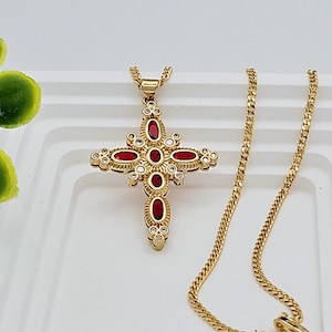 May include: A gold chain necklace with a red and white cross pendant. The cross is decorated with small white stones and red oval-shaped stones.