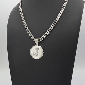 May include: A silver-toned curb chain necklace featuring a circular pendant with a scalloped edge. The center of the pendant displays a textured letter J. The metal has a matte, brushed finish throughout the entire piece, including the chain links.