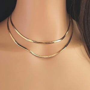 May include: Two gold chain necklaces layered on a person's neck. The chains are thin and have a delicate, slightly textured design.