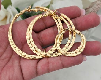 May include: Set of three gold-colored hoop earrings with a hammered texture. The earrings vary in size, with the largest hoop being approximately 2.5 inches in diameter. The earrings are displayed against a soft background.